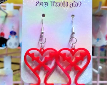 Pode incluir: Brincos vermelhos em forma de coração com um design de estrela, pendurados em ganchos prateados. Os brincos são exibidos em um cartão que diz "Pop Twilight". O fundo está desfocado, mostrando outras joias.
