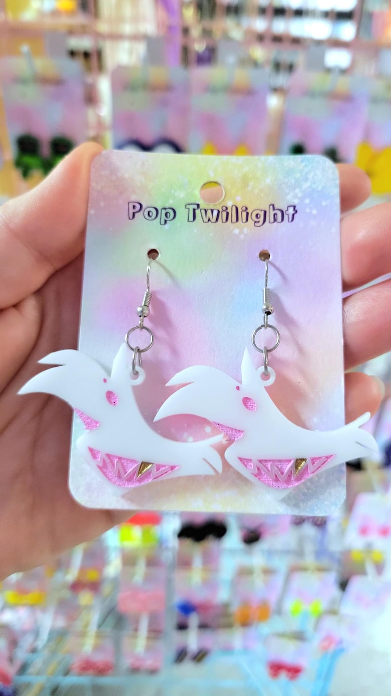 May include: White acrylic earrings with a bird-like design and pink and gold accents. The earrings hang from silver-colored hooks and are displayed on a card that reads "Pop Twilight".
