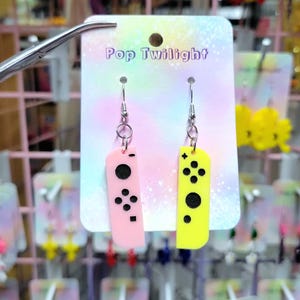 May include: Pink and yellow controller-shaped earrings with black button details. The earrings are displayed on a card that reads "Pop Twilight". Other earrings are visible in the background.