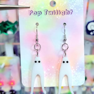 Puede incluir: Pendientes blancos con forma de fantasma y ojos de puntos negros, colgados de ganchos plateados. Los pendientes se exhiben en una tarjeta con el texto "Pop Twilight" en letras moradas sobre un fondo pastel. Un accesorio divertido y caprichoso.