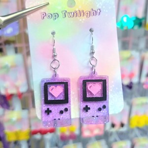 May include: A pair of purple glitter earrings shaped like retro handheld game consoles, each featuring a pixelated pink heart on the screen. The earrings are suspended from silver hooks and are displayed on a card that reads "Pop Twilight".