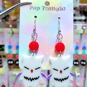 Puede incluir: Un par de pendientes Pop Twilight. Cada pendiente presenta un dije blanco con forma de fantasma con boca dentada, detalles morados y un punto rojo. Un círculo rojo y herrajes plateados completan el diseño.