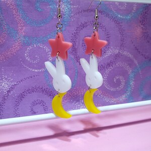 Puede incluir: Un par de pendientes colgantes con una estrella rosa, un conejito blanco y una luna creciente amarilla. Los pendientes cuelgan de ganchos para la oreja de color plateado. El fondo es una tela morada con un dise&ntilde;o de remolino.