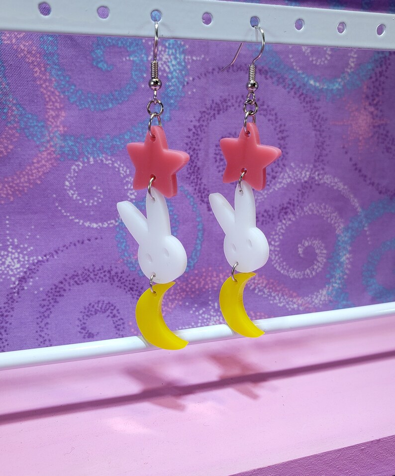 Puede incluir: Un par de pendientes colgantes con una estrella rosa, un conejito blanco y una luna creciente amarilla. Los pendientes cuelgan de ganchos para la oreja de color plateado. El fondo es una tela morada con un dise&ntilde;o de remolino.