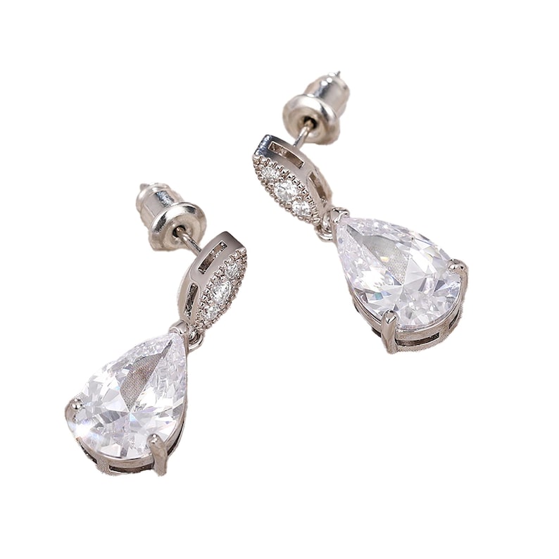 May include: A pair of silver-toned drop earrings. Each earring features a pear-shaped, clear gemstone suspended from a smaller, oval-shaped accent stone. The earrings have a post-and-clutch closure.