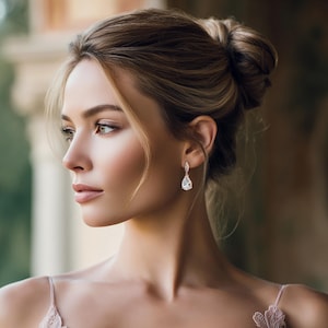 May include: A woman wearing elegant teardrop earrings with a sparkling, clear gemstone. The earrings are suspended from a delicate, silver-colored setting. The woman has her hair up in a bun and is wearing a light pink top.