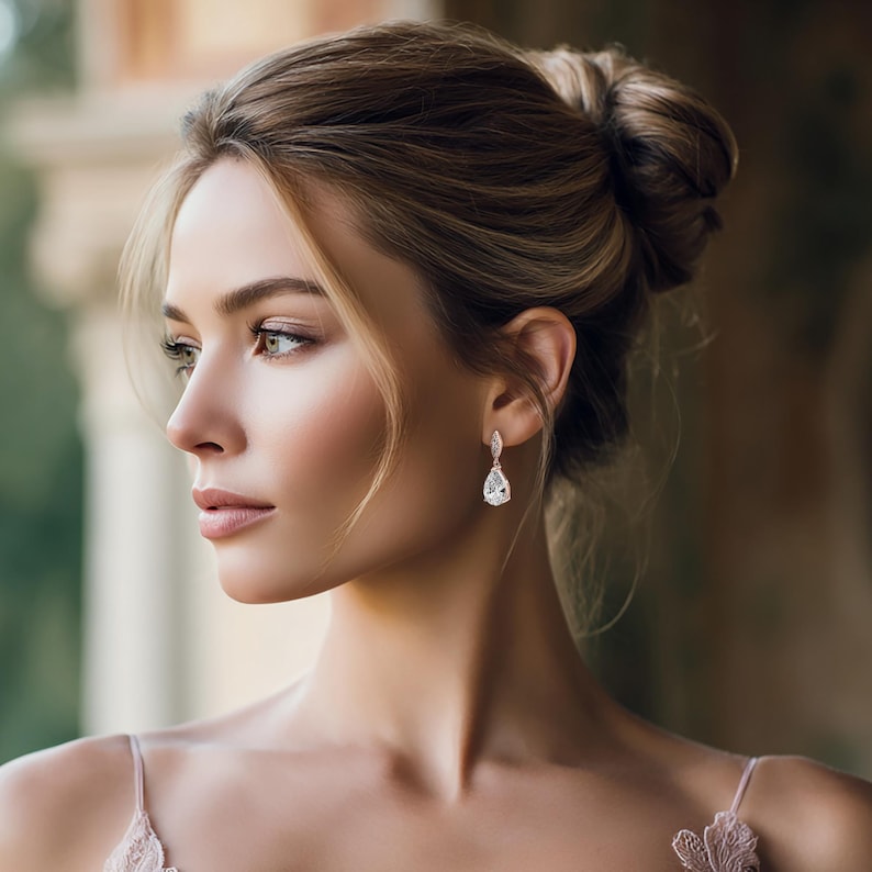 May include: A woman wearing elegant teardrop earrings with a sparkling, clear gemstone. The earrings are suspended from a delicate, silver-colored setting. The woman has her hair up in a bun and is wearing a light pink top.