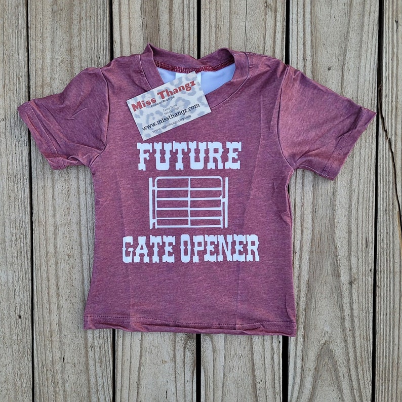 May include: A maroon short-sleeved t-shirt with the words "FUTURE GATE OPENER" in white block letters. A white gate graphic is centered between the words. A small tag is attached to the shirt.