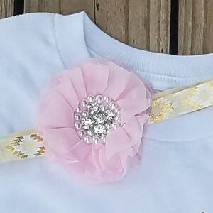 Pu&ograve; includere: Una maglietta bianca con un fiore rosa e un cerchietto con nastro dorato. Il fiore ha un centro di strass.