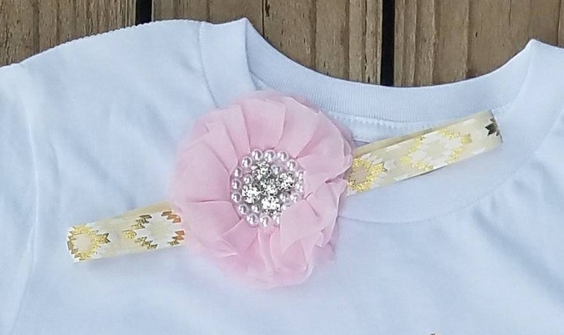 Pu&ograve; includere: Una maglietta bianca con un fiore rosa e un cerchietto con nastro dorato. Il fiore ha un centro di strass.