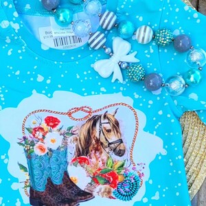 Puede incluir: Una camiseta azul turquesa con un patr&oacute;n de salpicaduras blancas. La camiseta presenta un gr&aacute;fico de un caballo marr&oacute;n con una bota vaquera azul y blanca y flores. La etiqueta de precio dice "Boo 19,99 &euro; Edad 5-6M 95% Algod&oacute;n 5% Spandex 33500636".