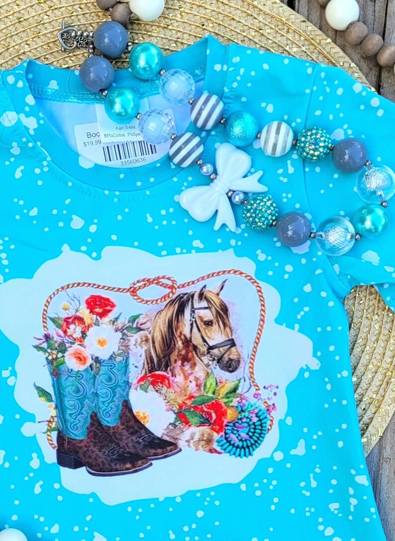 Puede incluir: Una camiseta azul turquesa con un patr&oacute;n de salpicaduras blancas. La camiseta presenta un gr&aacute;fico de un caballo marr&oacute;n con una bota vaquera azul y blanca y flores. La etiqueta de precio dice "Boo 19,99 &euro; Edad 5-6M 95% Algod&oacute;n 5% Spandex 33500636".