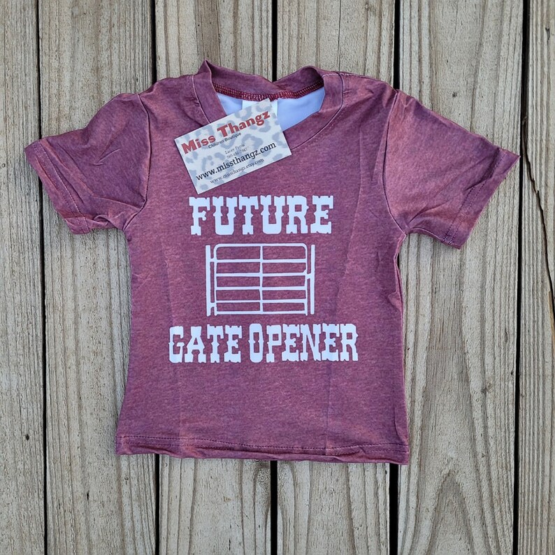 May include: A maroon short-sleeved t-shirt with the words "FUTURE GATE OPENER" in white block letters. A white gate graphic is centered between the words. A small tag is attached to the shirt.