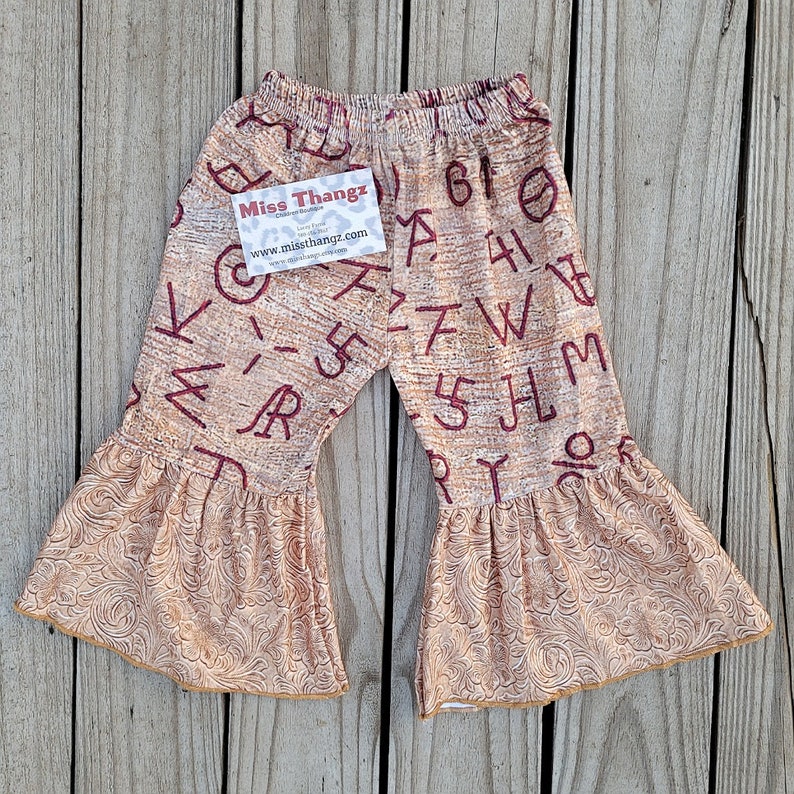 May include: Tan and burgundy bell-bottom pants with a western-themed pattern. The pants feature a repeating pattern of letters and numbers. The flared bottoms have a detailed, embossed design. A tag with the text "Miss Thangz" is attached.