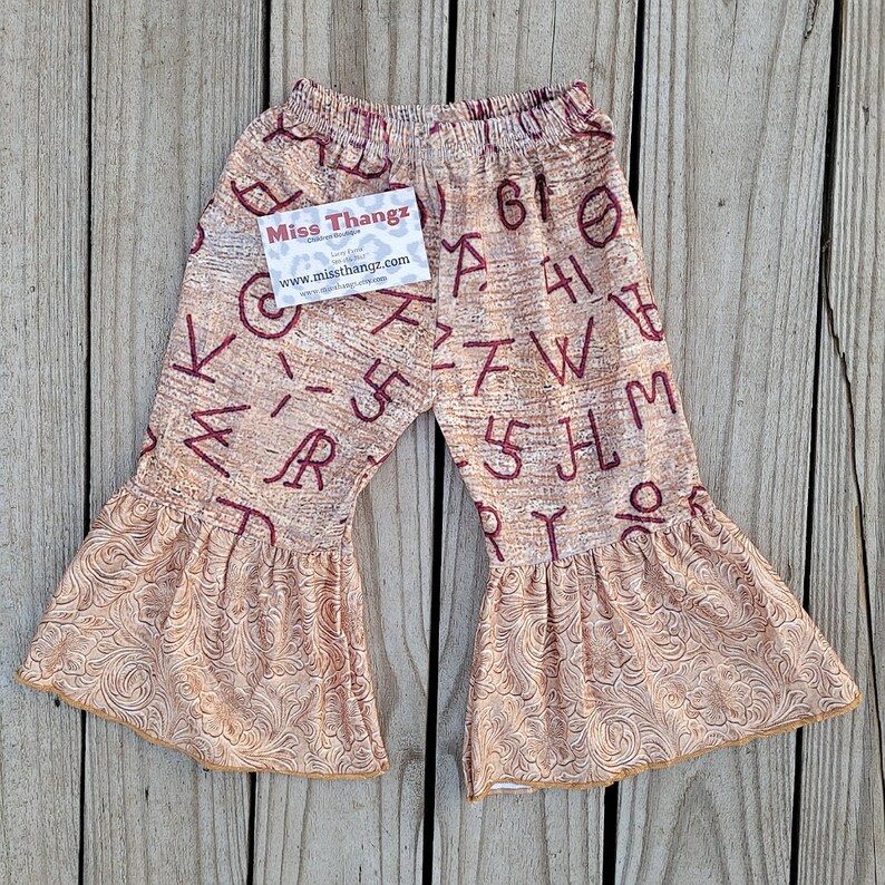 May include: Tan and burgundy bell-bottom pants with a western-themed pattern. The pants feature a repeating pattern of letters and numbers. The flared bottoms have a detailed, embossed design. A tag with the text "Miss Thangz" is attached.