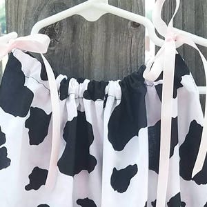 May include: A white and black cow print dress with pink ribbon straps hanging on a white hanger.