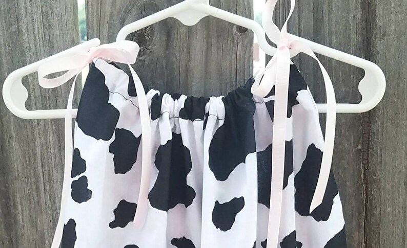 May include: A white and black cow print dress with pink ribbon straps hanging on a white hanger.