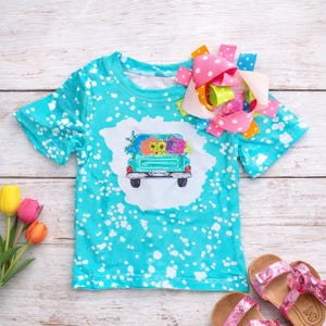 May include: A turquoise t-shirt with a white splattered pattern and a graphic of a teal pickup truck filled with colorful flowers. A large pink polka dot bow is attached to the shirt. Pink glitter sandals are visible.