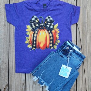 Pode incluir: Uma camiseta roxa de manga curta com um desenho de abóbora com um laço xadrez preto e branco. A abóbora é laranja e amarela, rodeada por folhas amarelas. Combinada com jeans azul.