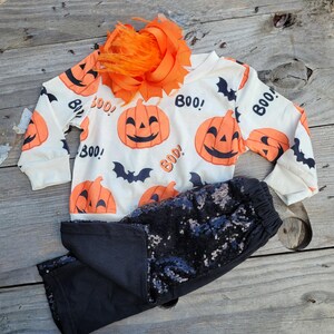 Peut inclure: Ensemble d'Halloween avec un haut à manches longues blanc orné de citrouilles oranges et du mot « Boo ! ». Le bas est un pantalon noir à paillettes, complété par un accessoire de cheveux orange.