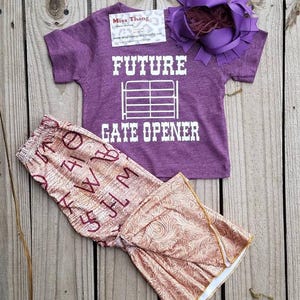 May include: A purple t-shirt with the text "FUTURE GATE OPENER" in white, paired with patterned pants and a purple bow. The pants have a floral design and the letters "W", "H", "M", and "5" printed on them. The bow has a feather accent.