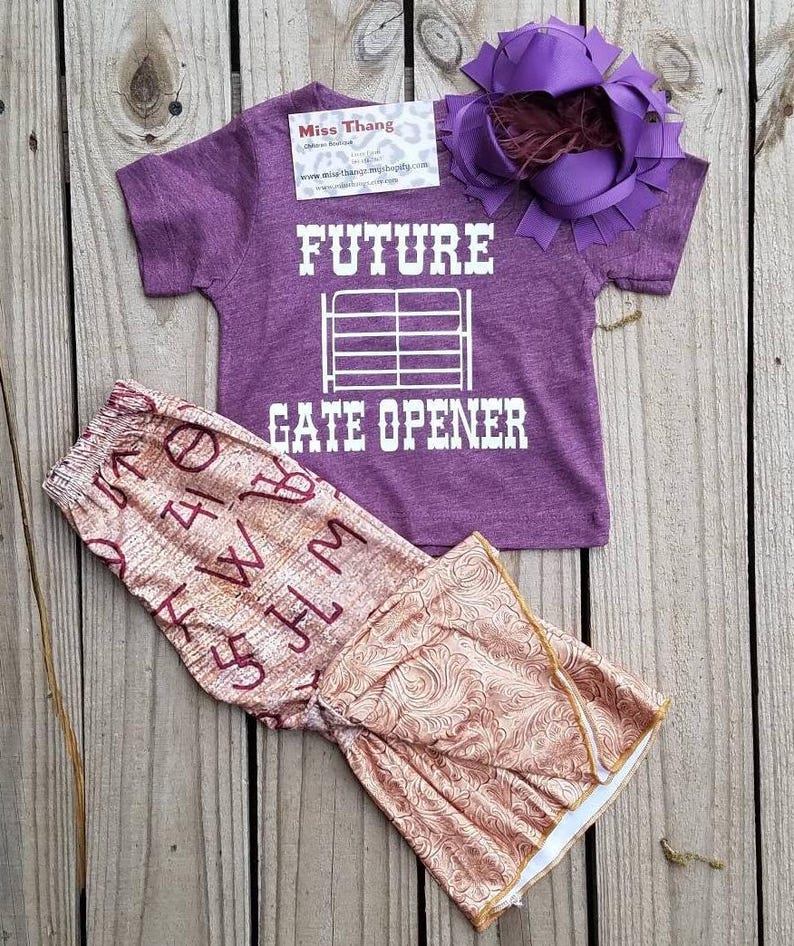 May include: A purple t-shirt with the text "FUTURE GATE OPENER" in white, paired with patterned pants and a purple bow. The pants have a floral design and the letters "W", "H", "M", and "5" printed on them. The bow has a feather accent.