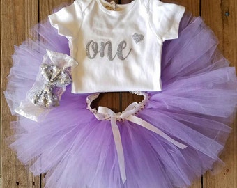 May include: A white baby bodysuit with the word "one" in silver glitter and a silver heart, paired with a lavender tutu skirt, a matching headband with a bow, and a white ribbon. The outfit is designed for a first birthday celebration.