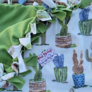 Könnte beinhalten: Eine grüne Fleece-Bindedecke mit weißen und braunen geknoteten Fransen. Die Decke hat einen weißen Hintergrund mit einem sich wiederholenden Muster von Kakteen in Töpfen.