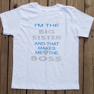 May include: White t-shirt with blue glitter text that reads "I'm the Big Sister and that makes me the Boss" with a blue glitter heart.