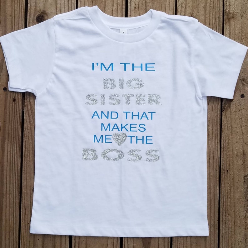 May include: White t-shirt with blue glitter text that reads "I'm the Big Sister and that makes me the Boss" with a blue glitter heart.