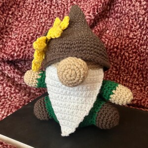 May include: A crocheted gnome with a brown hat, white beard, and green and brown clothing. The gnome has a yellow flower on its hat.
