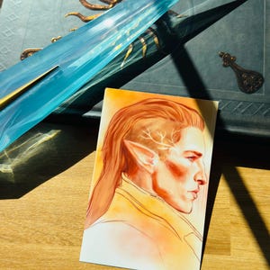 Puede incluir: Una ilustración de una persona con cabello largo rojo y orejas puntiagudas, de perfil, con una combinación de colores naranja claro y amarillo. Un objeto azul y dorado está en el fondo, junto con un libro azul oscuro con detalles dorados.