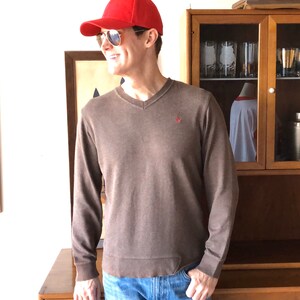 May include: A man wearing a brown long-sleeved sweater with a v-neck, a red baseball cap, and blue jeans. He is standing in front of a wooden cabinet with glass doors.