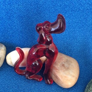 K&ouml;nnte beinhalten: Eine durchscheinende, rubinrote Hundefigur in sitzender Position. Der Hund hat einen gebogenen Schwanz und aufrechte Ohren. Er liegt auf einem glatten, hellfarbigen Stein vor blauem Hintergrund.
