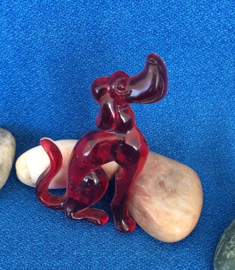K&ouml;nnte beinhalten: Eine durchscheinende, rubinrote Hundefigur in sitzender Position. Der Hund hat einen gebogenen Schwanz und aufrechte Ohren. Er liegt auf einem glatten, hellfarbigen Stein vor blauem Hintergrund.