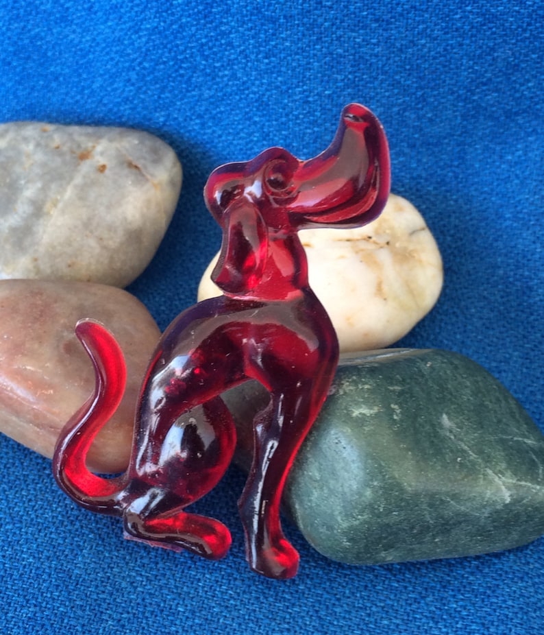 K&ouml;nnte beinhalten: Eine leuchtend rote Glasfigur eines Hundes in sitzender Position. Der Hund hat einen langen Schwanz und den Kopf erhoben. Die Figur steht vor einem blauen Hintergrund mit glatten Steinen.