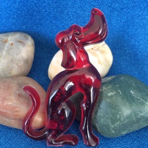 K&ouml;nnte beinhalten: Eine transparente, rubinrote Hundefigur mit erhobenem Kopf und eingerollter Rute. Der Hund sitzt, seine Beine sind sichtbar. Die Figur befindet sich vor einem blauen Hintergrund mit glatten, farbigen Steinen.