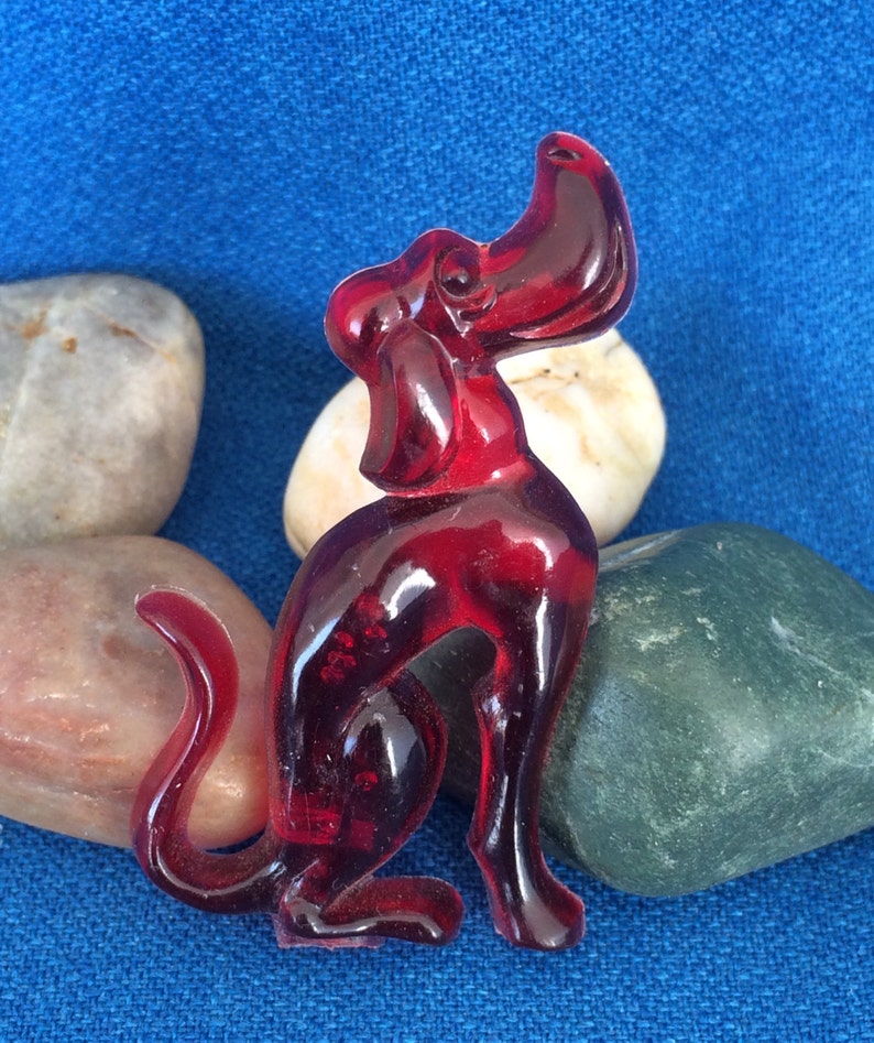 K&ouml;nnte beinhalten: Eine transparente, rubinrote Hundefigur mit erhobenem Kopf und eingerollter Rute. Der Hund sitzt, seine Beine sind sichtbar. Die Figur befindet sich vor einem blauen Hintergrund mit glatten, farbigen Steinen.