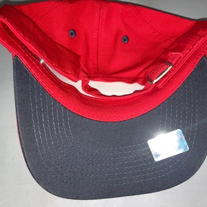 May include: A red baseball cap with a grey brim, viewed from the inside. The cap has a curved peak and an adjustable strap. The interior is red, and the peak is grey. A small sticker is visible on the peak.