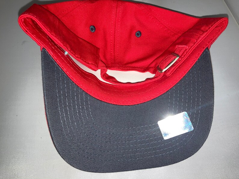 May include: A red baseball cap with a grey brim, viewed from the inside. The cap has a curved peak and an adjustable strap. The interior is red, and the peak is grey. A small sticker is visible on the peak.