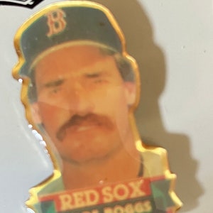 May include: A pin featuring a portrait of Wade Boggs, a baseball player for the Boston Red Sox. The pin has a gold border and includes the text "RED SOX WADE BOGGS" in red and green. Boggs is wearing a Red Sox cap.
