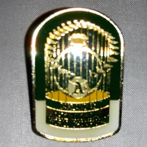 May include: A gold-colored baseball pin with a green background. The pin features a baseball stadium with a baseball player in the center. The pin has a white border.
