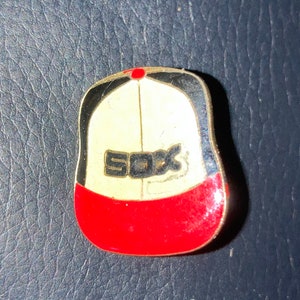 May include: A red, white, and black baseball cap pin with the word "SOX" in black letters on the front.