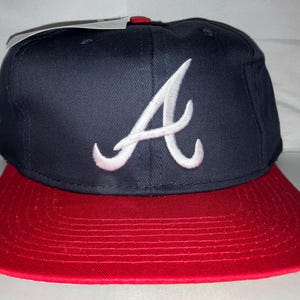 Op de afbeelding: Een marineblauwe baseballcap met een rode klep. De cap heeft een wit geborduurd "A" logo op de voorkant. De cap heeft een gestructureerde pasvorm en een platte klep.