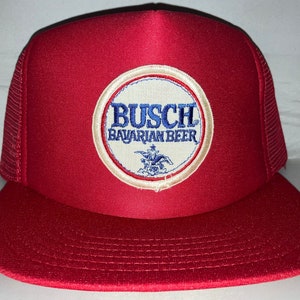 May include: Red and white trucker hat with a white circle patch that says "Busch Bavarian Beer" in blue lettering. The patch also features a blue and white eagle.