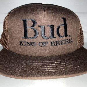 May include: A brown trucker hat with a curved brim. The front panel is brown with the word "Bud" embroidered in black, along with "KING OF BEERS." The mesh back is a lighter brown.