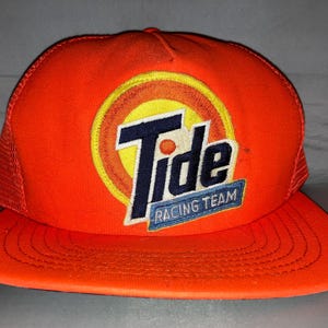Puede incluir: Una gorra de estilo camionero de color naranja brillante con parte trasera de malla y visera plana. El panel frontal muestra un logotipo circular bordado con un fondo de rayos de sol amarillos, el texto Tide en azul oscuro y un parche.