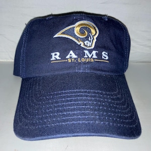 May include: A navy blue baseball cap with a white and gold embroidered St. Louis Rams logo. The logo features a ram's head with horns and the word "RAMS" written below it.