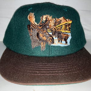 May include: A green and brown baseball cap with an embroidered image of a moose and other wildlife. The cap has a brown brim and a green crown.