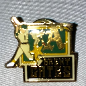 May include: A gold-colored pin featuring a cartoon baseball player in a green and yellow uniform, swinging a bat. The pin has a green and yellow background with a gold star and the text "BREW CITY CATTERS".